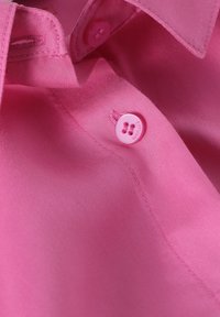 Helles rosa Stoffhemd mit glatter Textur, das eine Nahaufnahme eines Knopfes und des Kragens zeigt. Der Knopf ist in passendem hellrose und hat zwei Löcher.