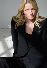 Szőke nő laza hajjal, fekete szabott blézert és nadrágot viselve, ülve, semleges szürke és fehér háttér előtt.