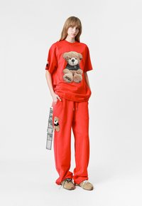 Camiseta roja de gran tamaño con gráfico de oso de peluche y pantalones de chándal a juego que presentan un parche de oso de peluche. Fabricada en tejido suave, con corte relajado y cintura con cordón.