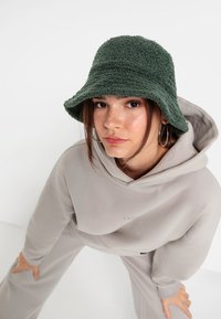 Cappello a secchiello verde e fluffoso con finitura testurizzata, indossato con una felpa grigia chiara. La felpa presenta un logo discreto sul davanti.