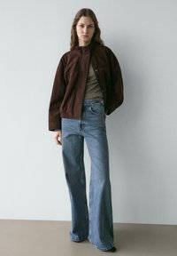 Braune Jacke aus Kunstwildleder mit hohem Kragen, kombiniert mit einem gestreiften Langarmshirt und weit geschnittenen blauen Jeans.