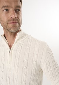 Cremfarbener Reißverschluss-Pullover mit einem Zopfmuster. Verfügt über einen hohen, gerippten Kragen und einen Reißverschluss am Hals. Weiche Textur.
