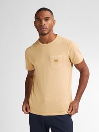 Tricou bej cu mânecă scurtă, realizat dintr-un material moale, având un buzunar pe piept cu o mică etichetă a brandului. Guler rotund și croială lejeră.