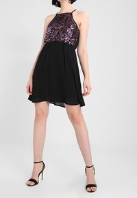 Kobieta w bezrękawnej czarnej sukience z fioletowym koronkowym topem i czarnych sandałach na wysokim obcasie, stojąca z nogami rozstawionymi na szarym tle.