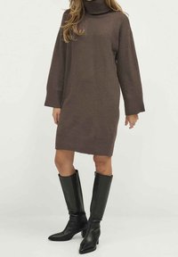 Kvinna som bär en brun långärmad polotröja klänning och svarta knähöga klackstövlar, stående på en vit bakgrund.