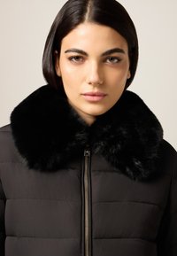 Schwarze Steppjacke mit einem weichen schwarzen Kunstpelzkragen. Verfügt über einen Reißverschluss und ein gestepptes Design, das eine glatte, matte Textur betont.