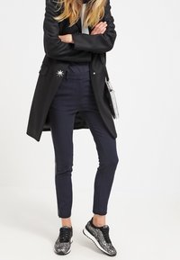 Černý kabát přes černý top, spárovaný s tmavě modrými slim-fit kalhotami a vzorovanými černými teniskami. Elegantní design a padnoucí silueta.