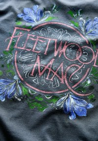 Fleetwood Mac Logo in pinker Schrift, umgeben von blauen und weißen Blumen auf grauem Stoffhintergrund.
