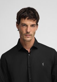 Camicia nera con collo a punta, maniche lunghe e apertura con bottoni. Presenta un piccolo logo ricamato sul lato sinistro del petto. Texture liscia.