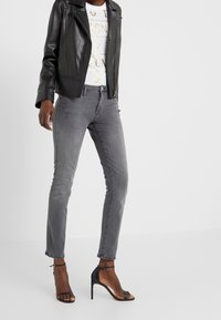 Osoba v černé kožené bundě, bílé tričko s potiskem, šedé skinny džíny a černé sandály na podpatku s kotníkovým páskem.