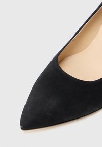 Sort ruskind sko med spids tåsæg med en glat overflade og eksponerede sider, der viser en neutral inderside, minimalt hardware og et slankt design.