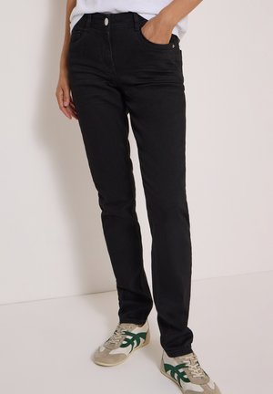 Personne portant un jean noir slim et des baskets blanches avec des bandes vertes, debout devant un fond clair uni.