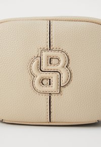 Beige teksturert lærveske med preget logo, sentral søm og kontrasterende sting. Firkantet form, glatt overflate og subtil glans.