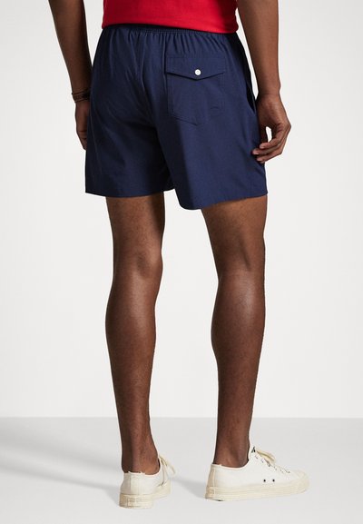 Ναυτρής μπλε μαγιό με ελαστική μέση, πίσω τσέπη με κουμπί και απλή υφή. Άσπρα αθλητικά παπούτσια συμπληρώνουν την εμφάνιση.