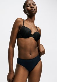 Γυναίκα που φοράει μαύρο σουτιέν με μπανέλα και σκουρόχρωμο ψηλοκομμένο εσώρουχο, στέκεται με το ένα χέρι στη μέση της μπροστά σε απλό φόντο.