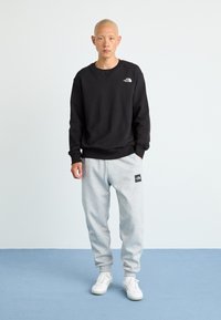 Svart hettegenser og grå joggebukser med et logo-patch, med en avslappet passform. Hvite sneakers fullfører antrekket mot en lys bakgrunn.