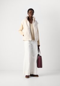 Gilet in finta pelliccia beige, top a maniche lunghe crema, pantaloni bianchi larghi e scarpe marroni. La modella tiene una borsa bordeaux. Design minimalista.