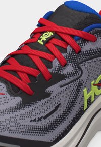 Αθλητικό παπούτσι με γκρι και μαύρο σχέδιο, φωτεινά κόκκινα κορδόνια, μπλε επένδυση στον αστράγαλο και νέον κίτρινο λογότυπο "HOKA" στη γλώσσα και την πλευρά.