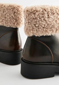 Brown leather ankle boots with a textured beige faux fur cuff, featuring a chunky black rubber sole and stitched detailing.
