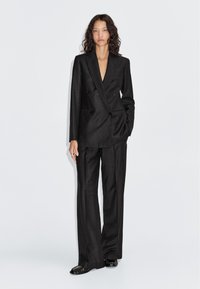 Zwart gestreept dubbelgeknoopt kostuum met broek met wijd uitlopende pijpen en een V-hals. De stof lijkt glad; voorzien van subtiele knopen en een getailleerde pasvorm.