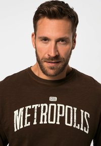 T-shirt marrone con la scritta "STEEL METROPOLIS" in grande con un effetto usurato in bianco. Collo tondo, maniche corte, texture morbida.