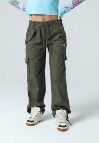 Olivgröna cargobyxor med dragsko i midjan, två framfickor, dekorativ broderi och vida ben, tillsammans med beigefärgade högsneakers.