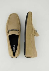 Beige Wildleder-Läufer mit einem eleganten Design, schwarzer Gummisohle, kontrastierendem Stich und einem dekorativen metallischen Akzent auf dem Obermaterial.