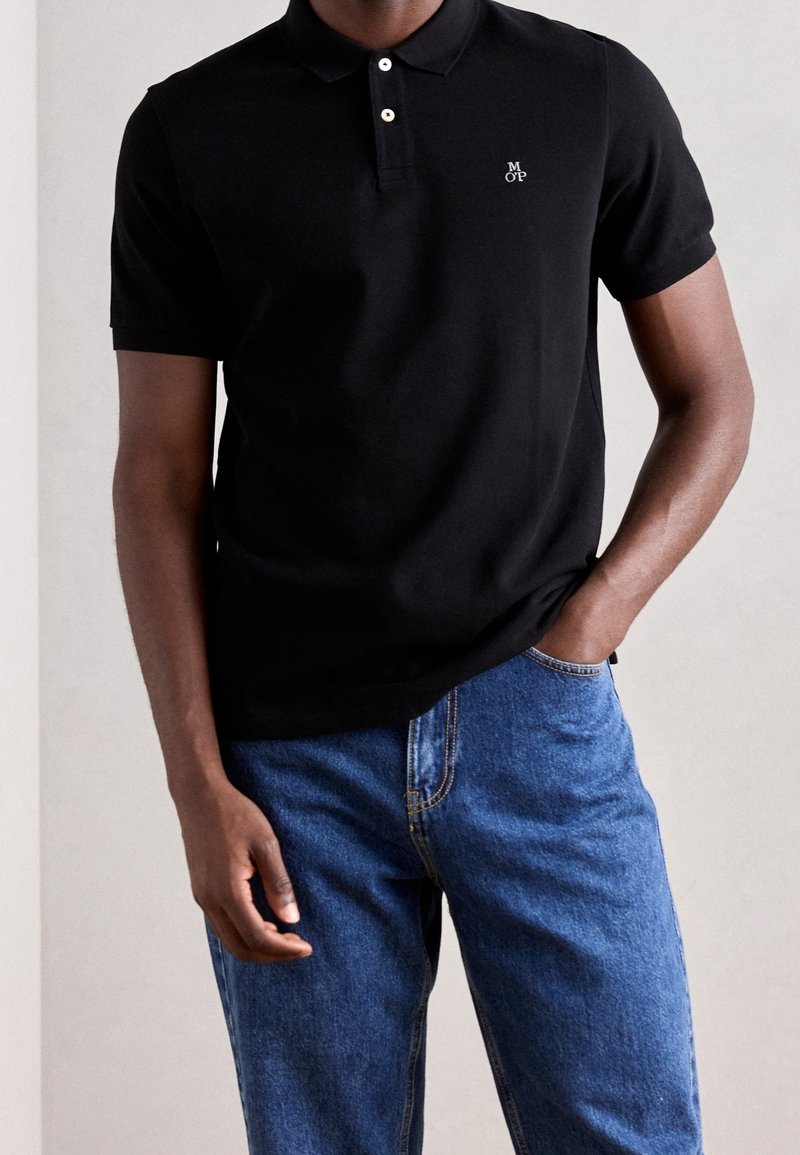 Polo noir avec une fermeture à trois boutons et un logo brodé, associé à un jean en denim bleu, offrant une coupe décontractée et une légère texture.
