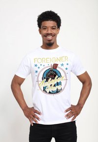 Bílý tričko s textem "FOREIGNER", grafikou jukeboxu a hvězdami v modré a zlaté barvě. Tričko má krátké rukávy a kulatý výstřih.