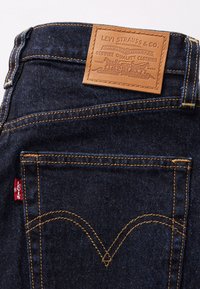 Temno modri denim kavbojkasti žep z oranžnim šivom, rdeča etiketka Levi's in svetlo rjav usnjen znak blagovne znamke Levi Strauss & Co. nad pasom.