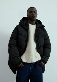 Svart puffjacka med huva, med ett quiltat mönster. Under, en krämfärgad ulltröja och blå denimbyxor. Enkel, modern stil.