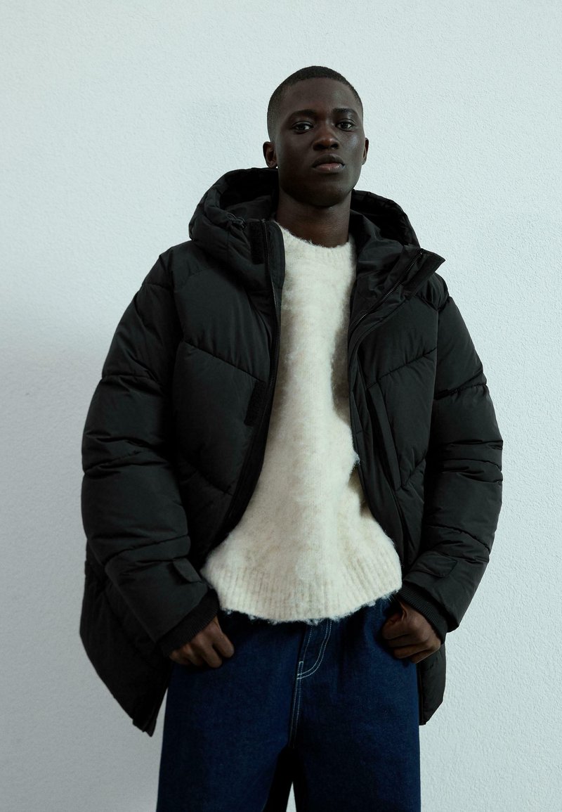 Svart puffjacka med huva, med ett quiltat mönster. Under, en krämfärgad ulltröja och blå denimbyxor. Enkel, modern stil.