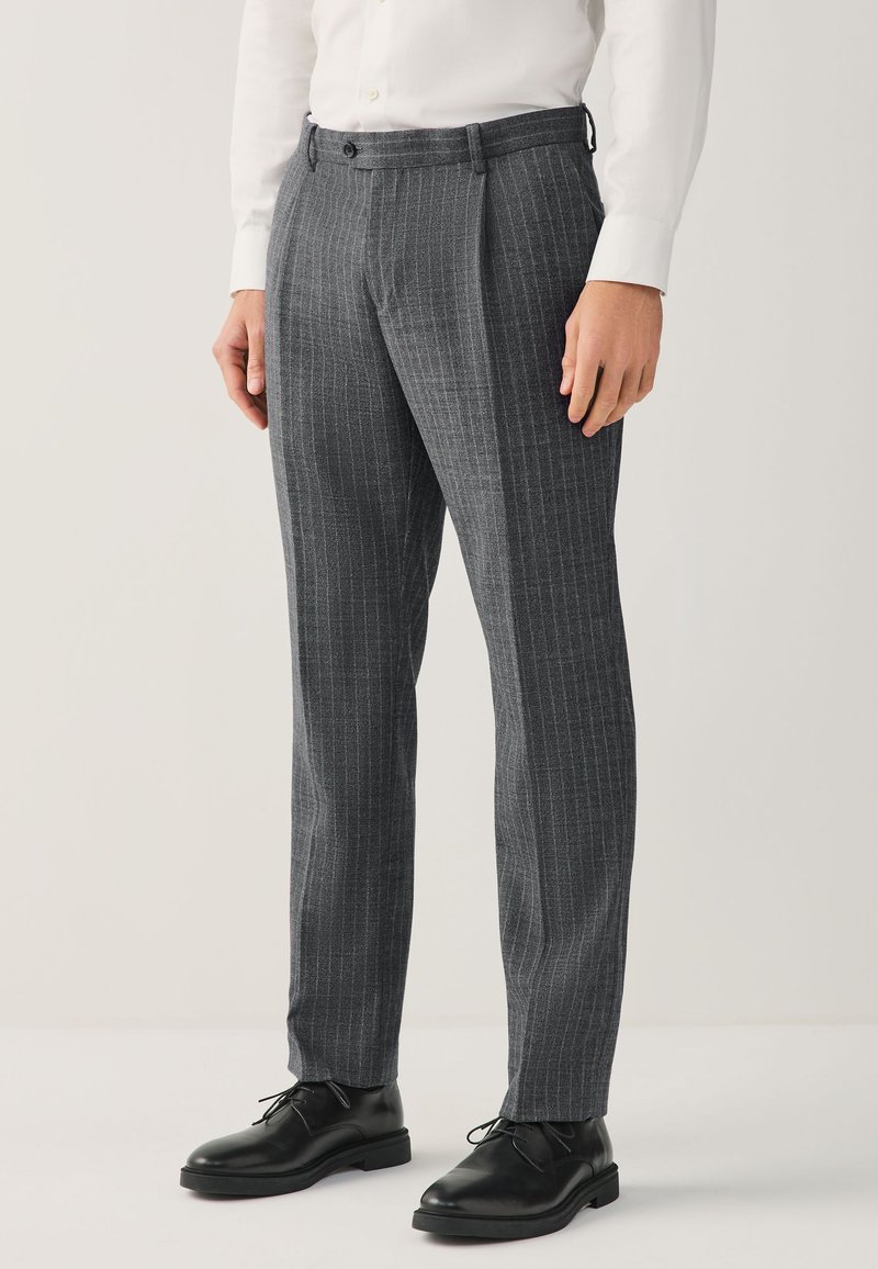 Homme portant un pantalon de costume gris à fines rayures, des chaussures en cuir noires et une chemise blanche, debout devant un fond neutre.