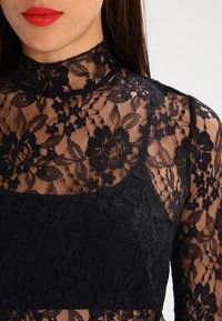 Top in pizzo nero con scollatura alta. Motivo floreale trasparente con dettagli testurizzati. Tessuto nero opaco nella zona del petto per copertura.