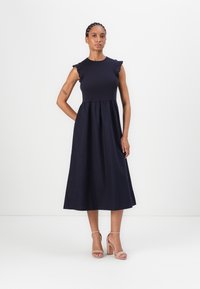 Navyblaues Midi-Kleid mit kurzen Rüschensäumen, einer taillierten Brustpartie und einem ausgestellten Rock. Getragen mit beigen Blockabsatzsandalen.
