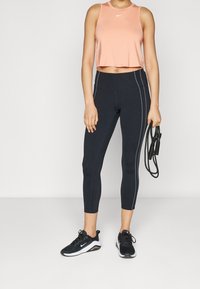 Ljusrosa kort sportlinne med rund hals och vit logotyp, kombinerad med svarta caprilegins med vita sidosömmar och sportskor.