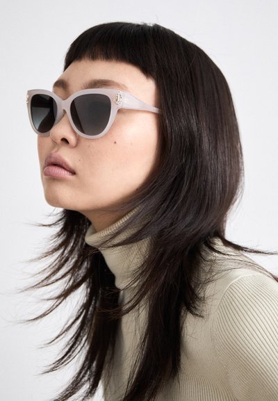 Lunettes de soleil grises en forme d'œil de chat avec des verres foncés. Monture transparente avec des accents en métal doré. Le mannequin a les cheveux droits et foncés et porte un col roulé à côtes.