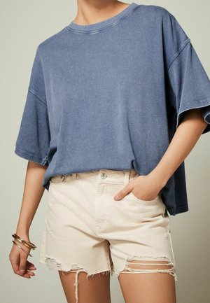 Person wearing a loose blue short-sleeve top and light beige distressed denim shorts, with bracelets on left wrist and right hand in pocket.
