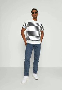 Camisa de mangas curtas listada em branco e azul-marinho com uma faixa sólida branca na barra, combinada com jeans azuis e ténis brancos, accessoriza com óculos de sol.