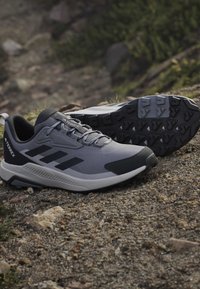 Trail running shoes in gray and black resting on rocky, uneven ground with visible rugged sole treads for outdoor use.