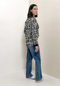 Schwarzer und weißer gemusterter Pullover mit Zickzackdesign, kombiniert mit blauen Denim-Weitbein-Hosen und weißen Sneakers. Neutraler Hintergrund.