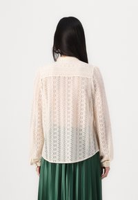 Blusa de encaje en color off-white con un patrón intrincado, mangas largas y un dobladillo curvo. Se lleva con una falda verde plisada para crear contraste.