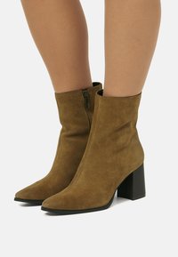 Frau trägt braune Wildleder-Stiefeletten mit Blockabsätzen und seitlichen Reißverschlüssen, die auf einer weißen Fläche stehen.
