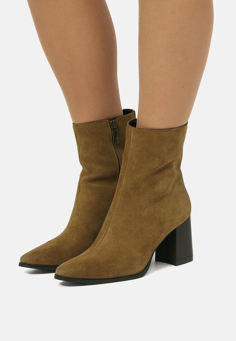 Donna che indossa stivaletti in suede marrone con tacco a blocco e cerniere laterali, in piedi su una superficie bianca.