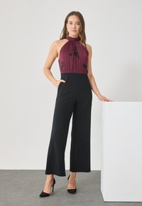 Jumpsuit bordeaux à dos nu avec un corsage plissé et un bas à jambes larges noir. Présente une broderie florale, des poches latérales et des chaussures à talons élégants.