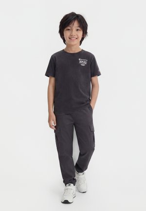 Niño de pie y sonriendo, vistiendo una camiseta negra con gráfico, pantalones cargo oscuros y zapatillas blancas sobre un fondo blanco liso.