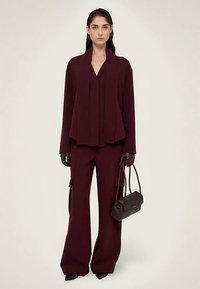 Completo giacca su misura bordeaux e pantaloni a gamba larga, con tessuto liscio e tasche cargo, abbinato a una piccola borsa nera e guanti.