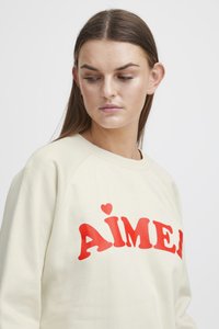 Cream-colored sweatshirt with raglan sleeves featuring a bold red "AÎMÉ" print and a small heart detail. Smooth texture, casual style.