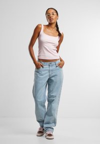 Hellrosa, taillierte Tanktop, kombiniert mit hochgürtigen, lockeren hellblauen Jeans und braunen Sneakers mit rosa Akzenten; das Model steht mit Händen in den Taschen.
