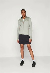 Svetlozelena zip jakna s temnim ovratnikom, s poudarki na oranžni zadrgi, v kombinaciji z modrimi kratkimi hlačami in črnimi športnimi copati.